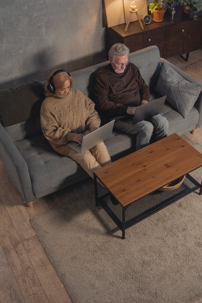 Elderly couple sitting on a sofa, using laptops for online activities in a cozy home setting.